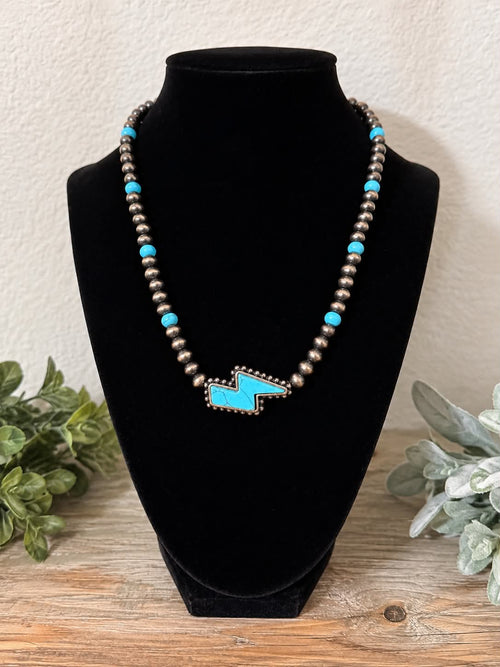 A beaded necklace displayed on a black velvet jewelry bust, featuring turquoise blue beads, metallic silver beads, and a turquoise and silver geometric pendant, set against a rustic wood and textured wall background.